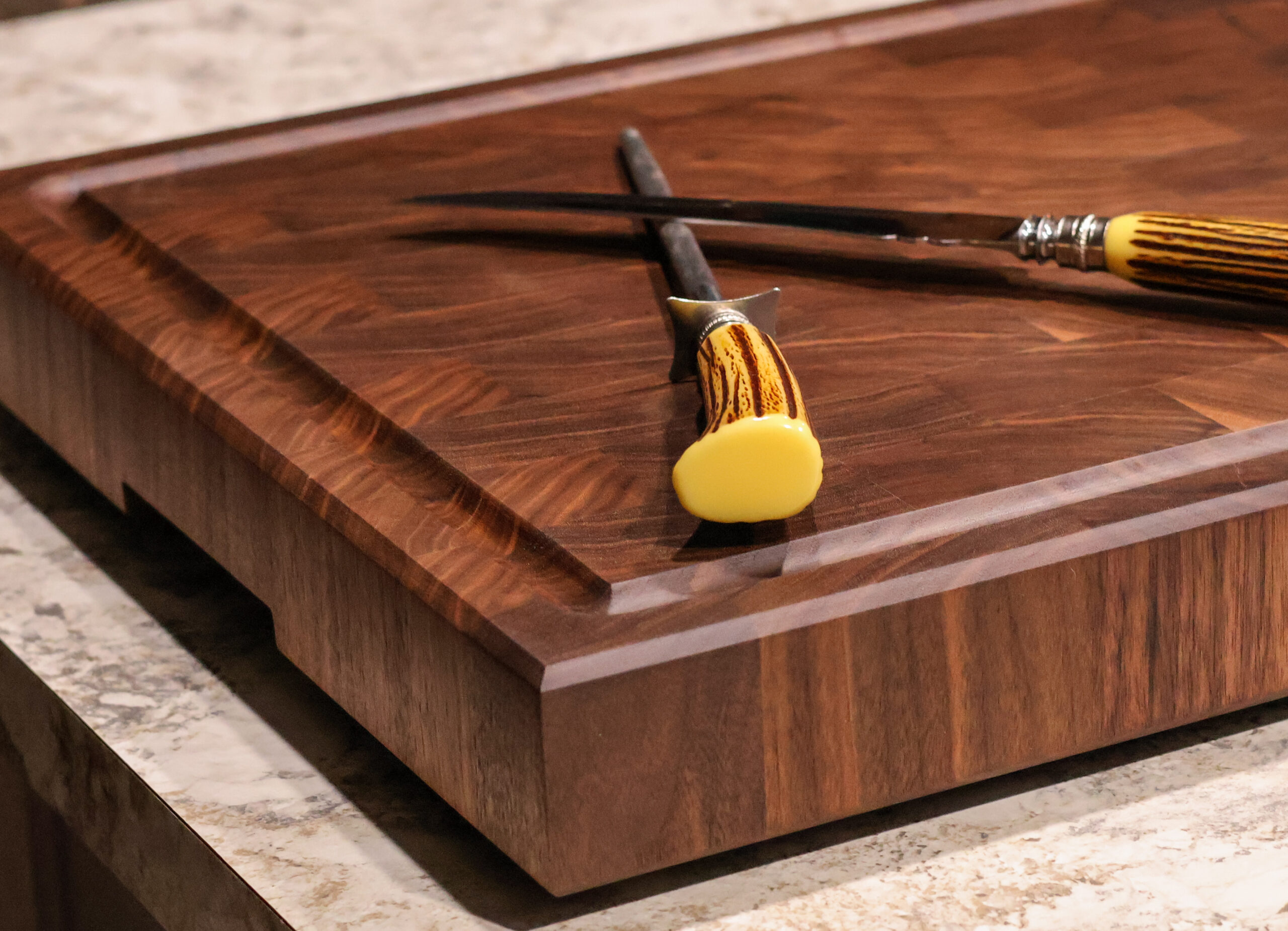 Close-up of a handcrafted walnut end-grain cutting board with deep juice groove, shown on a stone countertop with carving tools resting on the board's surface